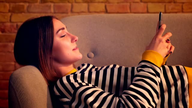 Closeup-side-view-portrait-of-young-pretty-girl-using-the-phone-and-smiling-happily-lying-on-the-couch-in-a-cozy-apartment-indoors