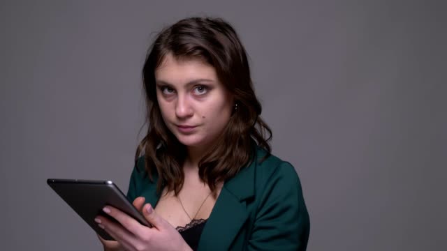 Closeup-shoot-of-adult-attractive-brunette-female-texting-on-the-tablet-and-looking-at-camera-with-background-isolated-on-gray