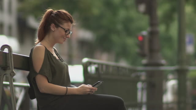 Cute-smiling-redhead-girl-with-glasses,-freckles,-piercings-and-red-hair-writing-a-text-message-on-her-smartphone-sitting-on-street-bench,-during-sunny-summer-in-Paris.-Slow-motion.-Trendy.