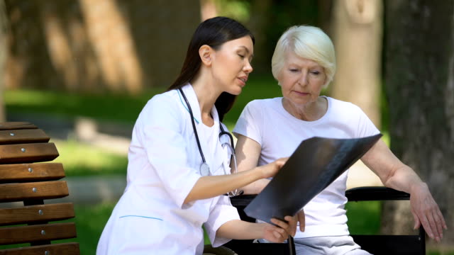 Lächelnde-Krankenschwester-zeigt-Spinal-Röntgen-zu-alten-Frau-im-Rollstuhl-im-Park,-Erholung