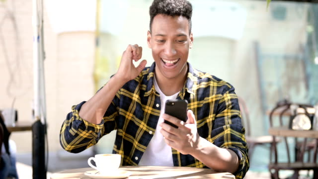 Young-African-Man-Celebrating-Success-on-Smartphone,-Outdoor-Cafe