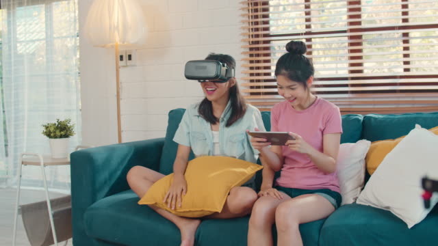Joven-pareja-lesbiana-asiática-usando-tableta-y-realidad-virtual-jugando-juegos-juntos-mientras-se-acuesta-sofá-en-la-sala-de-estar-en-casa.