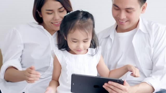 Asiatische-Familie-sitzt-auf-dem-Bett-lachend-und-suchen-in-digitalen-Tablet,-während-im-Schlafzimmer-liegen