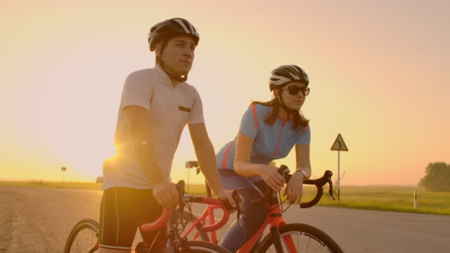 Ein-Mann-und-eine-Frau-in-Helmen-mit-Fahrrädern-stehen-und-reden-bei-Sonnenuntergang.-Ruhe-nach-einer-Radtour-auf-der-Autobahn.-Rennräder.-Paar-Sport.