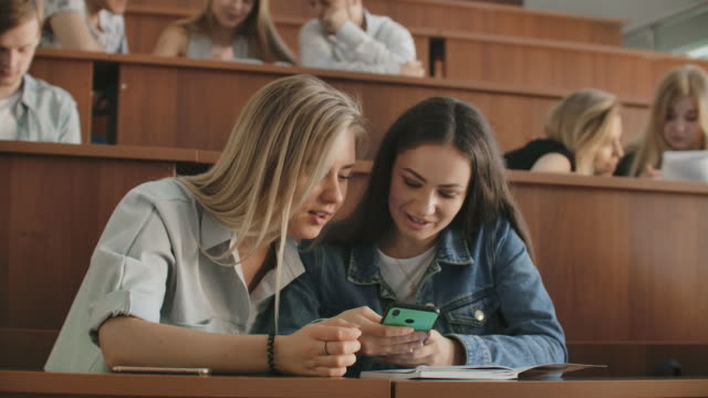 Chicas-bonitas-estudiantes-están-utilizando-el-teléfono-inteligente,-viendo-la-pantalla,-hablando-y-riendo-sentado-en-los-escritorios-de-la-universidad.-Redes-sociales,-internet,-millennials-y-concepto-educativo.
