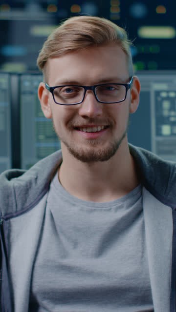 Porträt-eines-intelligenten-und-hübschen-IT-Spezialisten,-der-brillen-trägt,-lächelt-vor-der-Kamera.-Im-Hintergrund-Personal-Computer-mit-Bildschirmen-im-Rechenzentrum.-Vertikale-Bildschirmausrichtung-Video-9:16