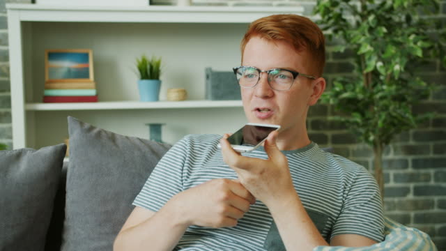 Joyful-young-man-recording-audio-message-using-smart-phone-in-apartment