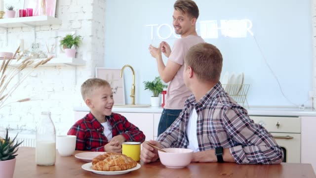 Glückliche-schwule-Familie-zwei-Väter-und-Sohn-frühstücken-in-der-Küche.
