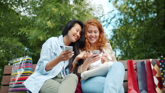 Joyful-friends-using-smartphones-outdoors-in-park-laughing-discussing-content