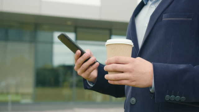 A-businessman-stands-outside-the-business-center-with-a-Cup-of-coffee-in-his-hand.-He's-texting-on-his-smartphone.-He's-wearing-a-suit-and-glasses.-Close-up-shooting.-4K