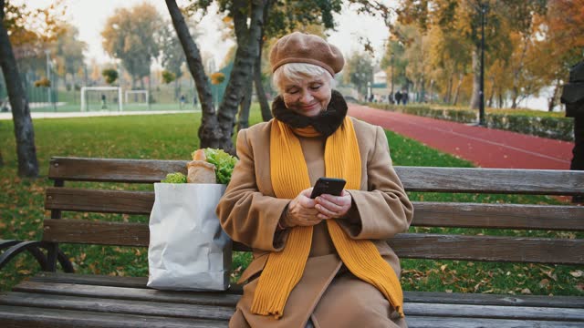 Ältere-Frauen-tippen-Nachricht-auf-Handy-sitzen-auf-der-Bank-im-Herbst-park,-Papiertüte-mit-Lebensmitteln-ist-in-der-Nähe