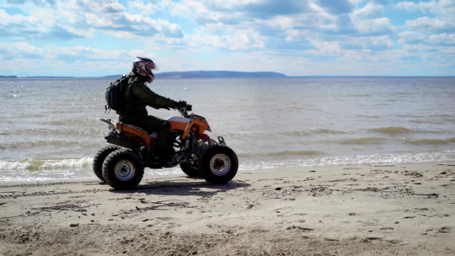 Mann-gekleidet-in-Schutzausrüstung-und-Helm-ATV-Reiten-und-fahren-entlang-der-Küste-mit-hoher-Geschwindigkeit.-Fan-von-Extremtouristen-mit-Rucksack-hinter-Rauschen-auf-seinem-Quad-Bike-am-Strand