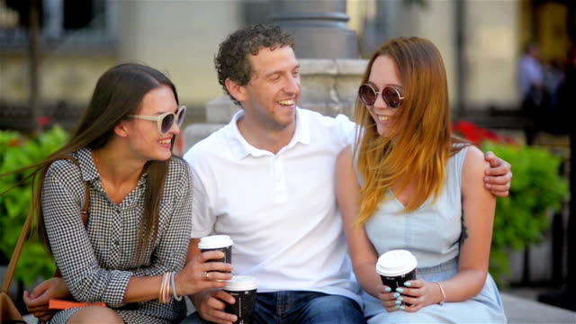 Portrait-of-Three-Friends-Drinking-Coffee-Sitting-on-the-Bench-Outdoors-in-the-City-During-Warm-Summer-Day.-Boy-is-Hugging-Girl-in-Light-Blue-Dress.-Another-Woman-Has-Long-Dark-Hair-and-Sunglasses