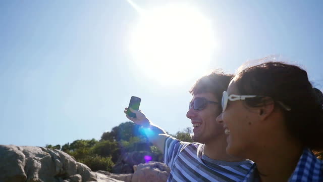 Young-smiling-couple-wearing-sunglasses-taking-selfie-portrait-by-beautiful-mountain-with-lense-flare-effects-on-the-sun-on-the-background-in-slow-motion.-1920x1080