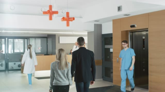 In-the-Hospital-Young-Couple-Walks-to-Reception.-Medical-Personnel,-Doctors,-Nurses,-Assistants-and-Patients-in-the-Busy-Lobby.-New,-Modern-Medical-Facility.