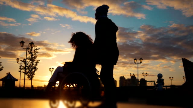 Caring-man-with-a-disabled-woman-in-wheelchair-walking-through-the-quay-at-sunset