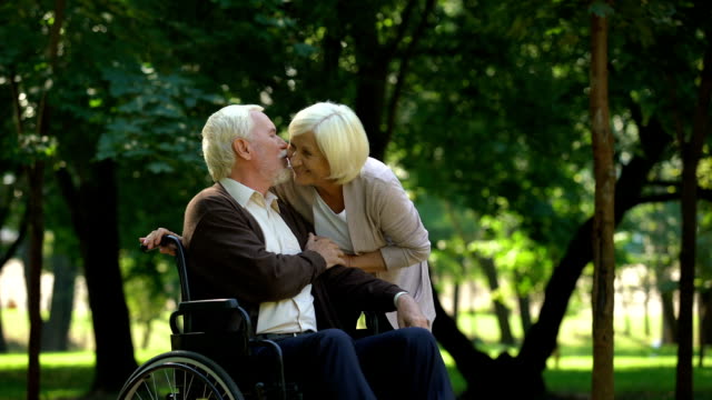 Mature-couple-resting-in-park-looking-at-grandkids,-man-sitting-in-wheelchair