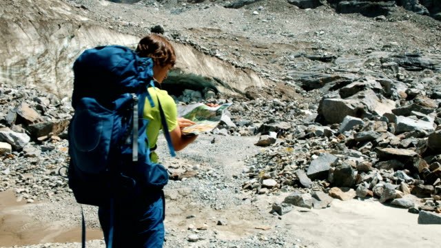 Turismo-hombre-camina-con-una-mochila,-estudia-un-mapa-y-muestra-una-ruta-en-las-montañas-contra-un-glaciar-azul,-lenta.