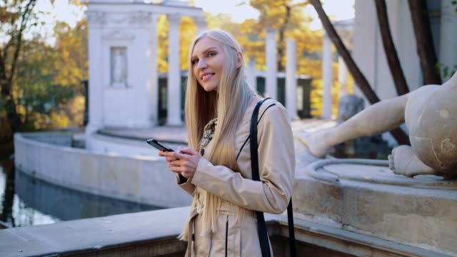 Mujer-rubia-alegre-en-la-capa-de-color-beige-en-el-parque-de-otoño,-mirando-su-smartphone