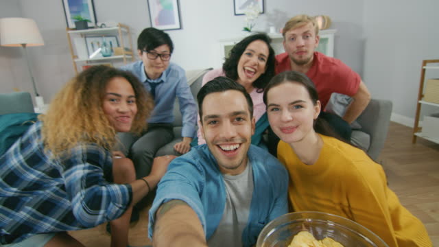 At-Home-Diverse-Group-of-Friends-Doing-Collective-Video-Stream,-Guy-Lifts-Camera.-People-Smile,-Laugh,-Wave-and-Greet-Viewers.Young-People-Doing-Live-Stream-of-the-Home-Party.