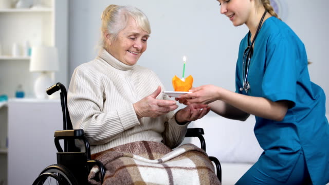 Doctor-giving-birthday-cake-to-handicapped-woman,-aged-lady-blowing-out-candle
