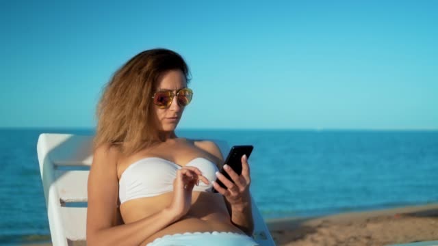 Young-Smiling-Mixed-Race-Tourist-Girl-in-Sunglasses-Using-Mobile-Phone-at-the-Beach-near-the-Sea-with-Beautiful-Sunset