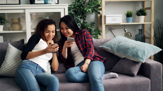 Cheerful-students-Asian-and-African-American-are-talking-and-laughing-looking-at-smartphone-screen-using-gadgets-sitting-on-sofa-at-home.-Youth-and-devices-concept.