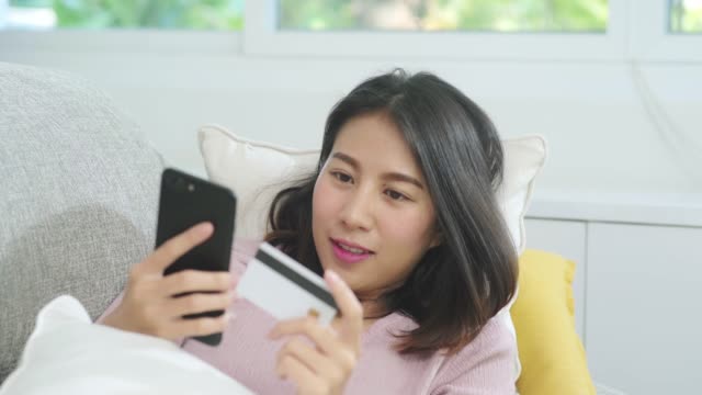 Young-smiling-Asian-woman-using-smartphone-buying-online-shopping-by-credit-card-while-lying-on-sofa-when-relax-in-living-room-at-home.-Lifestyle-latin-and-hispanic-ethnicity-women-at-house-concept.