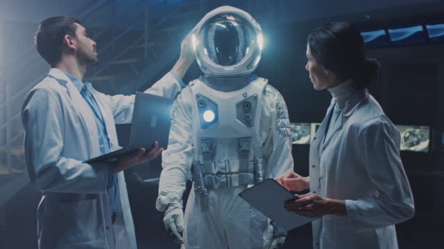 Diverso-equipo-de-científicos-aeroespaciales-e-ingenieros-que-llevan-abrigos-blancos-tienen-discusión,-usar-computadoras,-diseño-nuevo-traje-espacial-adaptado-para-la-exploración-y-el-viaje-Galaxy.-Construyendo-traje-de-astronauta