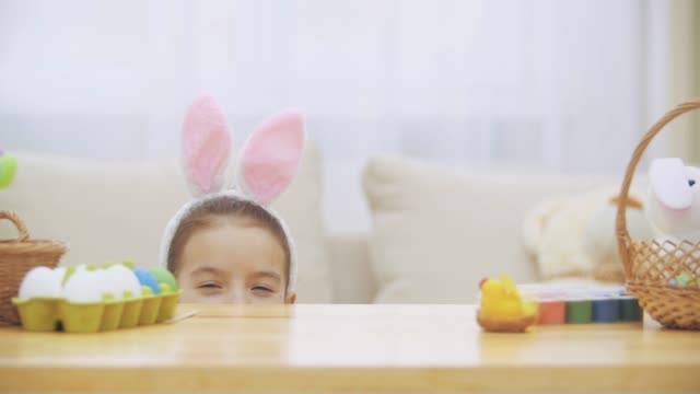 Cute-little-girl-with-bunny-ears-is-hiding-under-the-table-full-of-Easter-decorations.-Little-cute-white-girl-is-making-faces.-Laught-is-in-the-room.