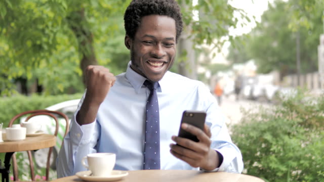 Hombre-de-negocios-africano-aclamación-éxito-en-Smartphone-sentado-en-al-aire-libre-Cafe