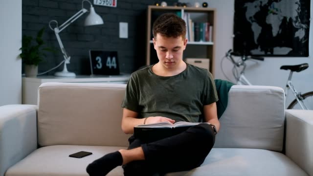 Ein-Junge-liest-aufmerksam-ein-Buch-auf-einer-Couch,-wenn-er-von-seinem-Handy-abgelenkt-wird-und-im-Internet-surfen