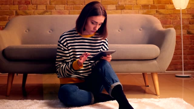 Closeup-portrait-of-young-pretty-girl-using-the-tablet-sitting-on-the-floor-in-a-cozy-apartment-indoors
