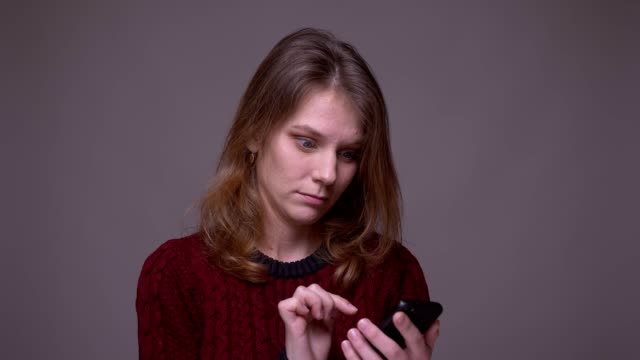 Junge-Studentin-wischt-Fotos-auf-dem-Smartphone-und-reagiert-emotional-auf-grauem-Hintergrund.