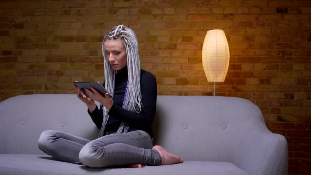 Closeup-shoot-of-young-attractive-caucasian-hipster-female-browsing-on-the-tablet-messaging-on-the-couch-indoors-in-a-cozy-apartment
