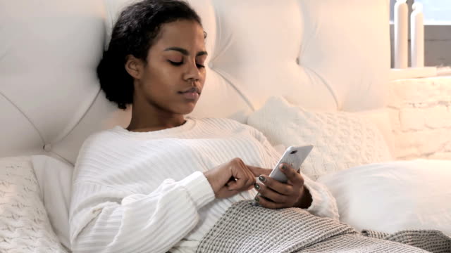 Junge-afrikanische-Frau-mit-Smartphone-beim-Entspannen-im-Bett