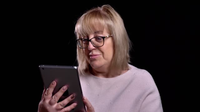 Closeup-shoot-of-old-caucasian-blonde-grandma-in-glasses-surfing-socail-media-on-the-tablet-in-front-of-the-camera-with-background-isolated-on-black