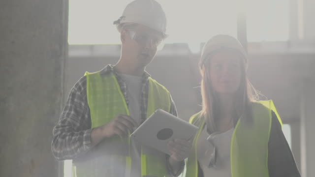 Bauarbeiter-Mann-und-Architekt-Frau-in-einem-Helm,-diskutieren-den-Plan-des-Baus-des-Hauses,-erzählen-einander-über-den-Entwurf,-halten-eine-Tablette,-Blick-auf-die-Zeichnungen,-Hintergrund-der-Sonnenstrahlen