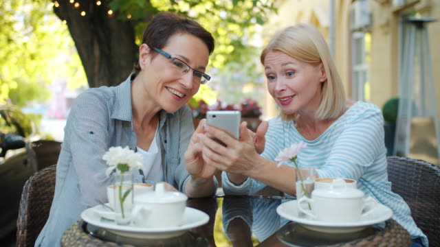 Excited-women-using-smartphone-laughing-in-open-air-cafe-in-city-street