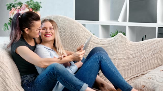 Zwei-glückliche-junge-charmante-lesbische-Mädchen-Blick-auf-Bildschirm-von-Tablet-PC-Umarmung-liegend-auf-der-Couch