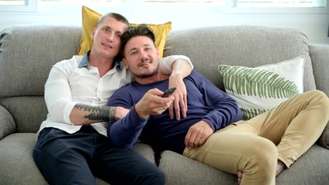 Schwules-Paar-entspannen-doneriert-auf-der-Couch-und-schaut-fern.-Switch-Kanal-und-sprechen.