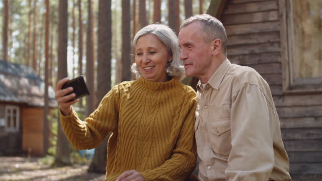 Pareja-senior-haciendo-selfie-cerca-de-wooden-Country-Cabin