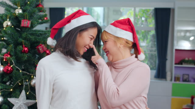 Pareja-lesbiana-asiática-celebran-festival-de-Navidad.-LGBTQ-adolescente-mujer-usan-sombrero-de-Navidad-relajarse-feliz-sonriendo-mirando-a-la-cámara-disfrutar-de-las-vacaciones-de-invierno-de-Navidad-juntos-en-la-sala-de-estar-en-casa.