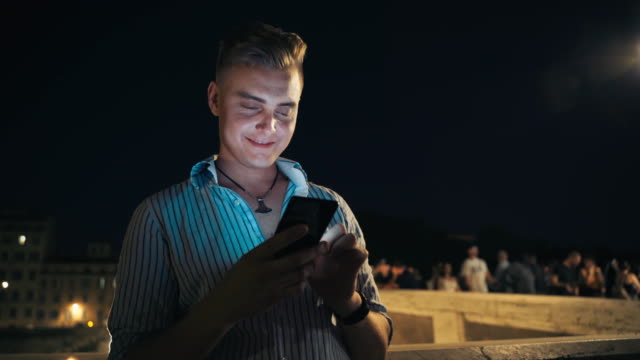 Schön-lächelnder-Mann,-der-nachts-geht-und-sein-Handy-benutzt.-Europäische-Touristen,-die-Daten-online-überprüfen-und-mit-Menschen-chatten,-die-mobile-Apps-nutzen.-Mann-auf-dem-Hintergrund-der-beleuchteten-Nacht-überfülltstraße.-Mann-mit-Telefon-Ch