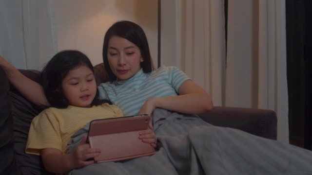 Junge-asiatische-Familie-und-Tochter-glücklich-mit-Tablet-zu-Hause.-Koreanische-Mutter-entspannen-mit-kleinen-Mädchen-beobachten-Film-auf-dem-Sofa-liegen,-bevor-sie-im-Wohnzimmer-im-Haus-in-der-Nacht-schlafen.