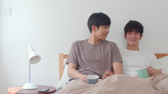 Asian-Gay-men-couple-talking-having-a-great-time-at-modern-home.-Young-Asia-lover-male-happy-relax-rest-drink-coffee-after-wake-up-while-lying-on-bed-in-bedroom-at-house-in-the-morning-concept.