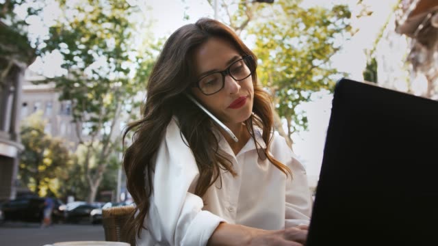 Lady-in-glasses,-white-shirt.-She-sitting-at-table-with-laptop-in-an-outdoor-cafe,-talking-on-smartphone-and-typing.-Business-concept.-Close-up