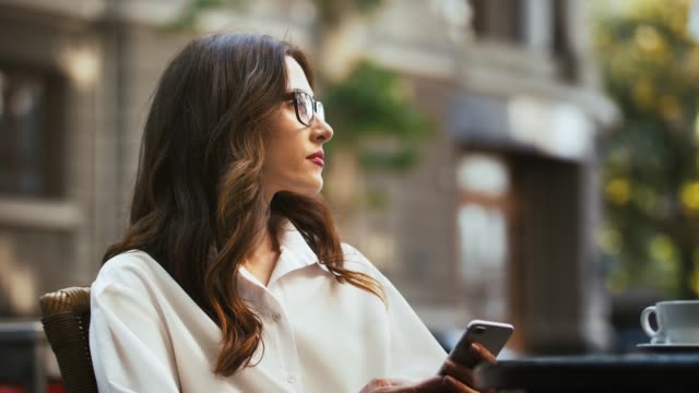 Business-woman-in-glasses,-white-shirt.-Sitting-at-table-with-cup-of-coffee-in-outdoor-cafe.-Browsing-news-on-cellphone,-looking-around.-Slow-motion