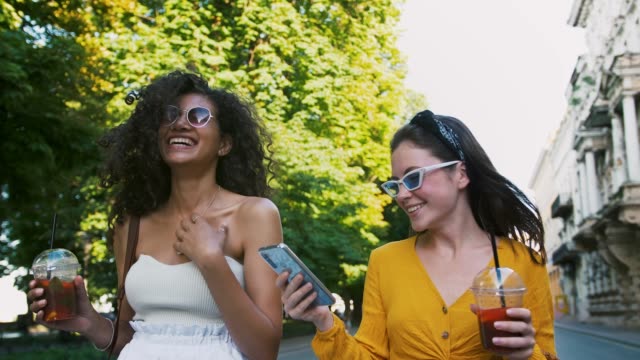 Girls-holding-cold-tea-in-plastic-cups.-Laughing,-looking-surprised-watching-video-on-mobile-phone.-Walking-along-city-avenue.-Close-up,-slow-motion
