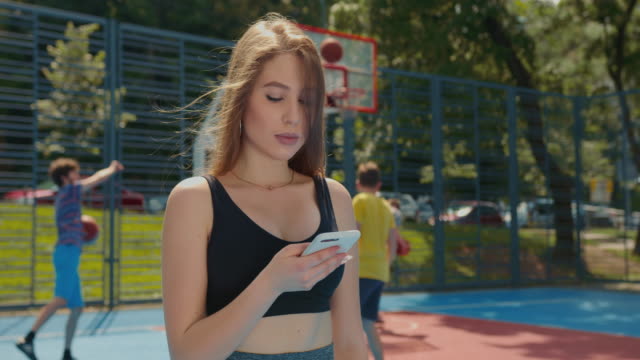 A-sporty-girl-is-texting-on-her-smartphone.-Her-hair-is-flying-in-the-wind.-She's-wearing-a-sports-uniform.-People-are-playing-floorball-in-the-background.-4K